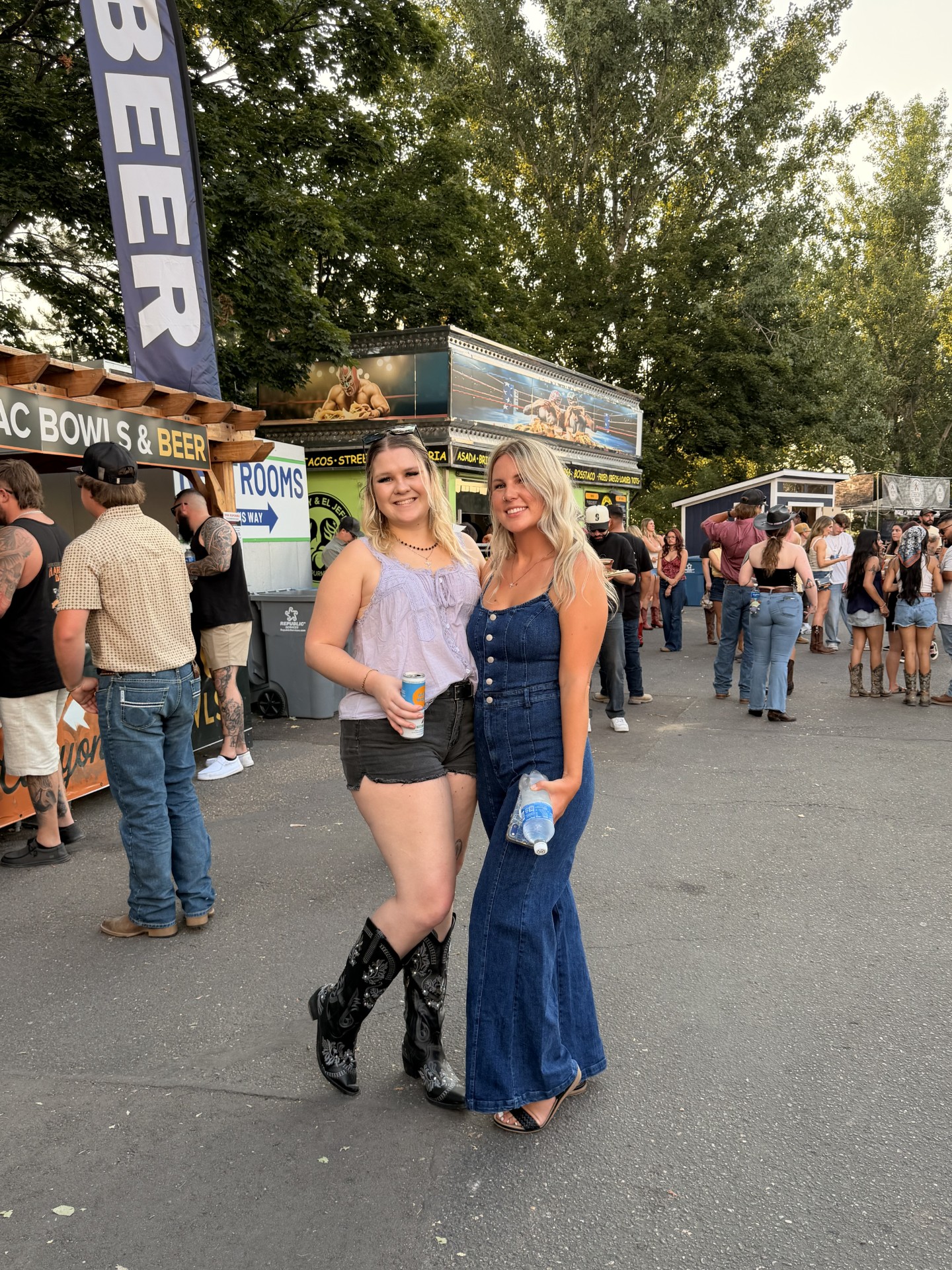 Two blondes at the Koe Wetzel concert standing near concession booths. The left one in black boots, shorts, and a lavender top. The right in a denim romper.